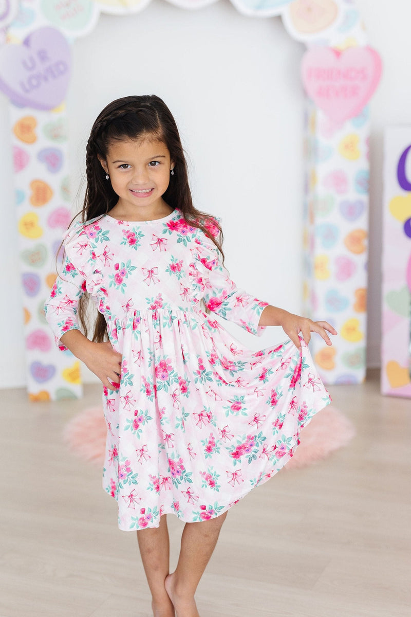 A little girl wearing the Gingham Bows 3/4 Sleeve Ruffle Twirl Dress, joyfully posing and twirling with a charming smile, surrounded by a playful backdrop of pastel decorations.