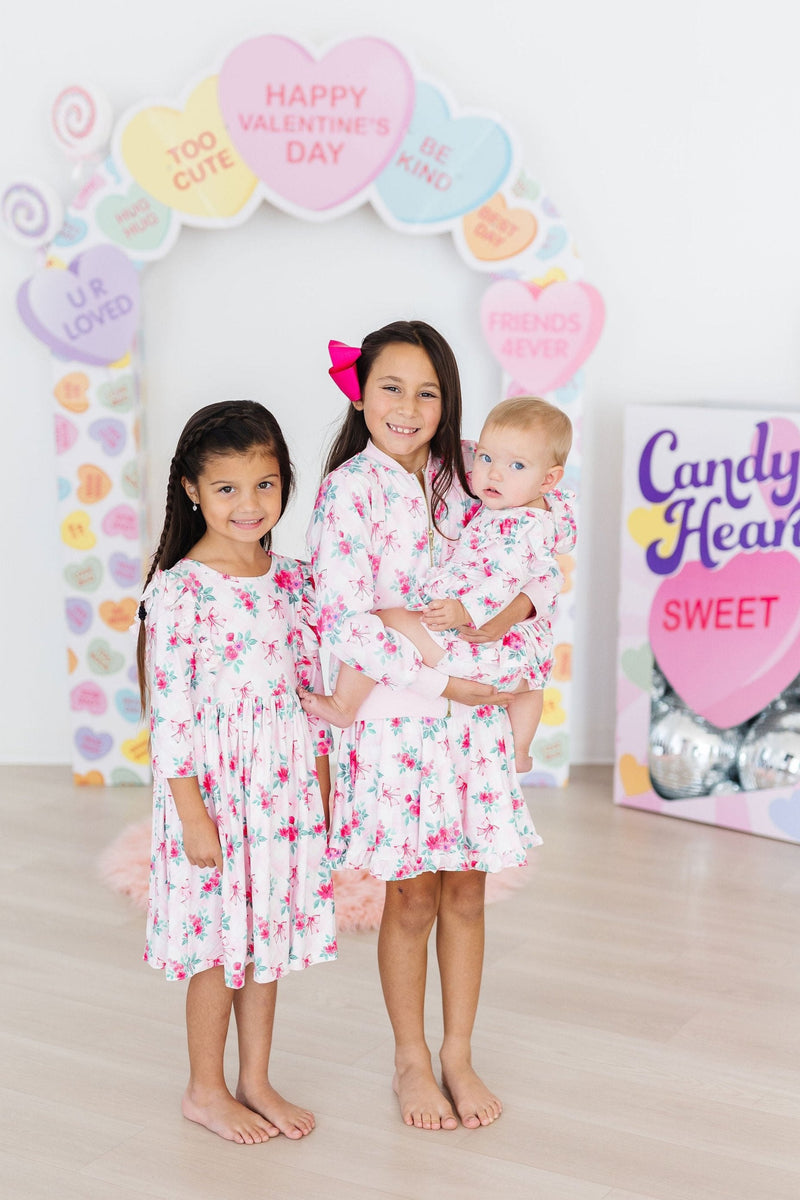 Three adorable girls styled in the Gingham Bows Satin Jacket by Mila & Rose, embracing friendship and fashion with vibrant floral dresses and radiant smiles, perfect for any playful occasion.