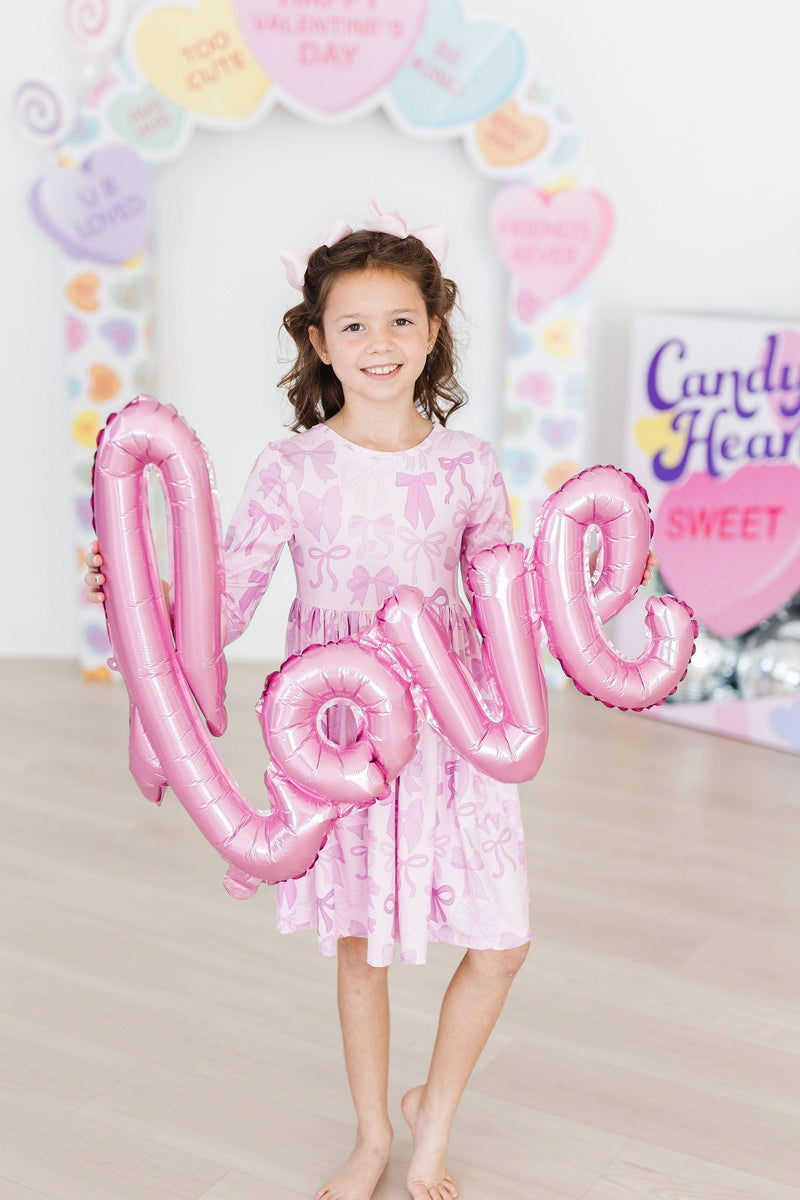 A joyful girl wearing the Twinkle Ties 3/4 Sleeve Pocket Twirl Dress from Mila & Rose, holding a pink 'love' balloon in a cheerful setting.