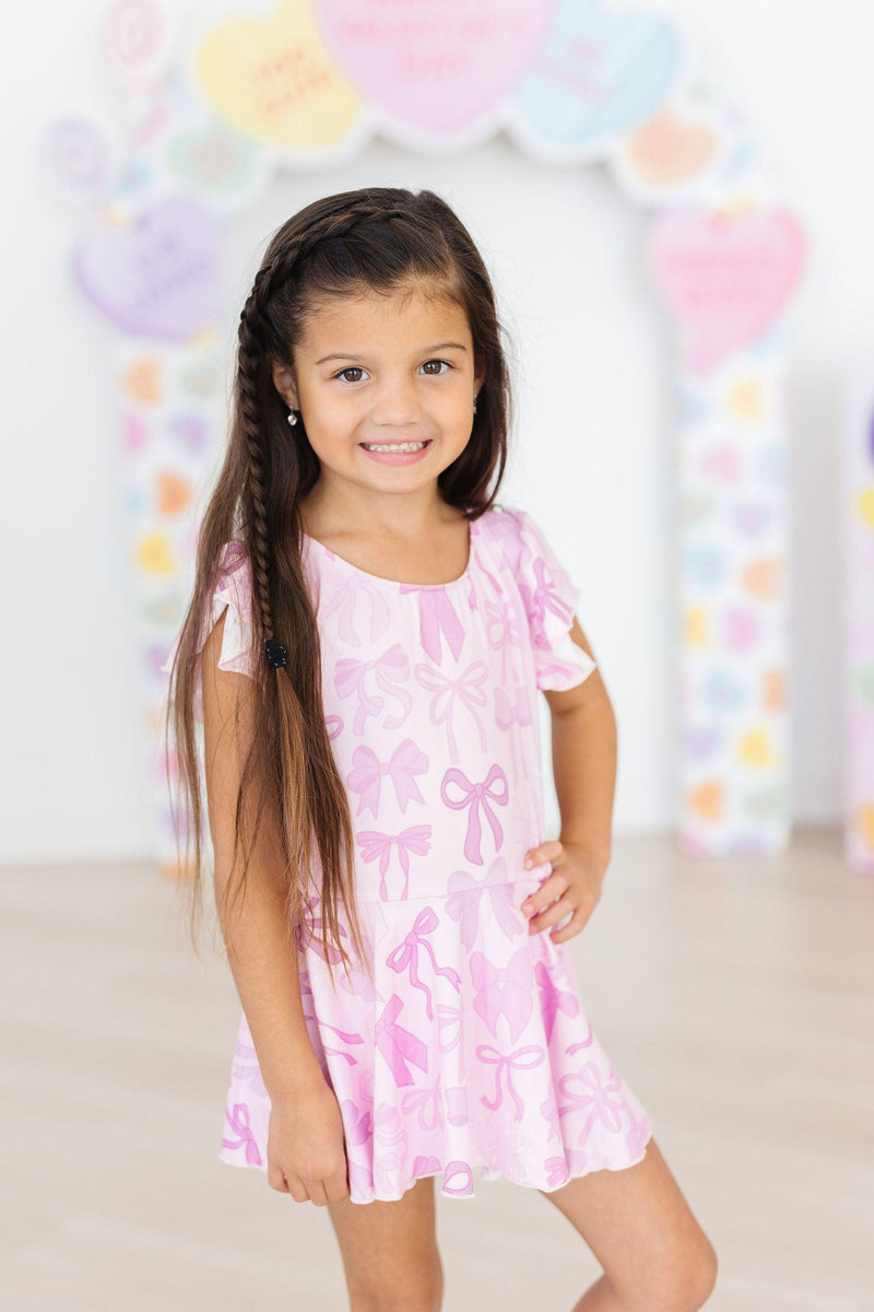 A girl in a pink Twinkle Ties S/S Twirl Leotard by Mila & Rose, confidently posing with a smile on her face, colorful decor for Valentine's Day in the background.