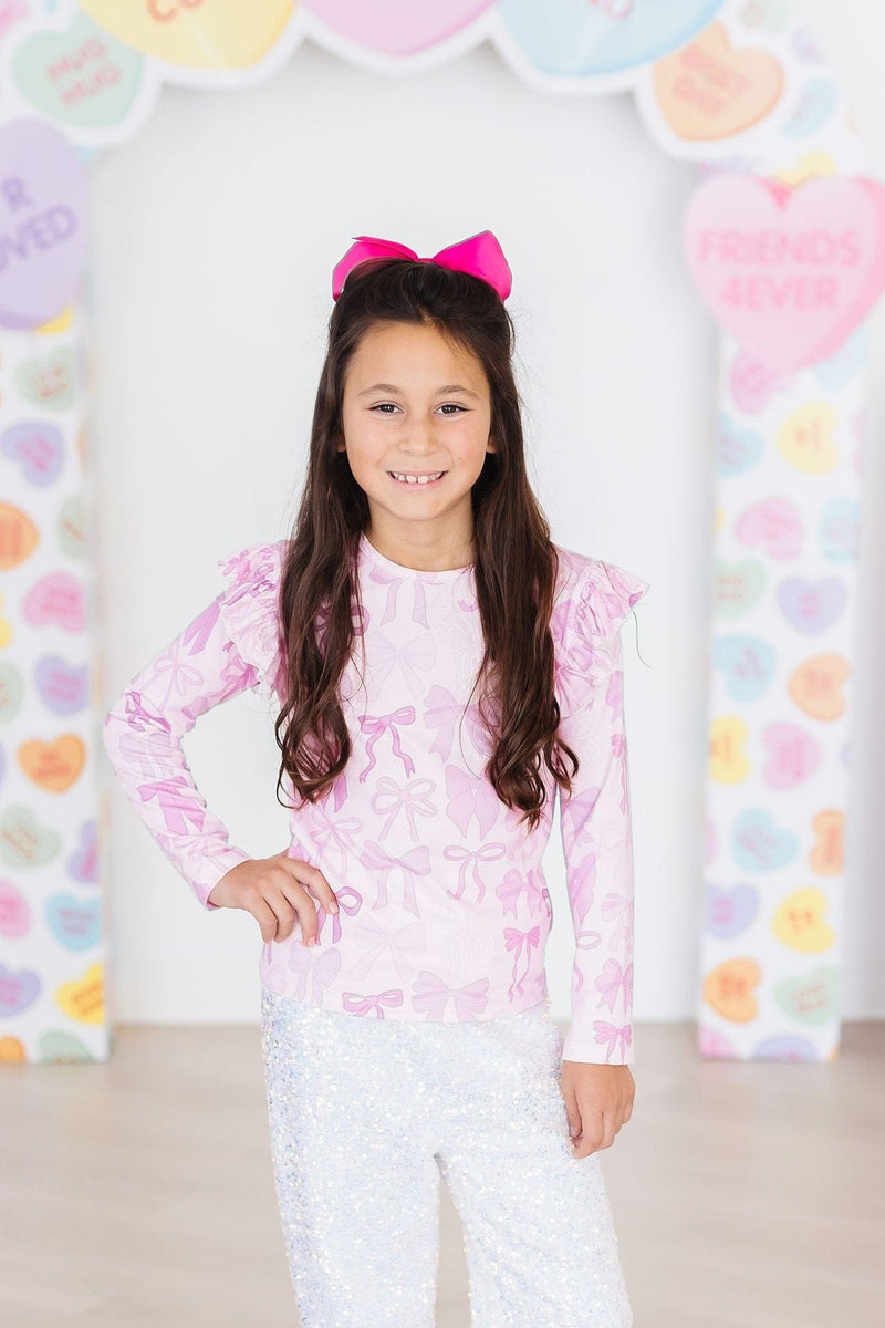 A happy girl wearing the Twinkle Ties L/S Ruffle Tee with pink bows, complemented by a large pink bow in her hair.