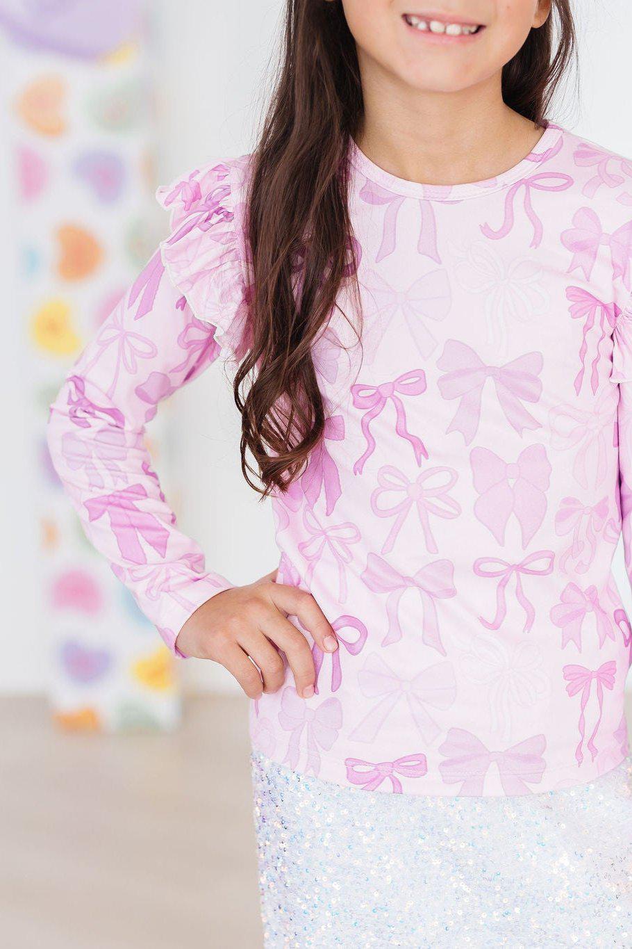 Twinkle Ties L/S Ruffle Tee styled by a smiling girl, featuring playful pink bow patterns and ruffled sleeves.