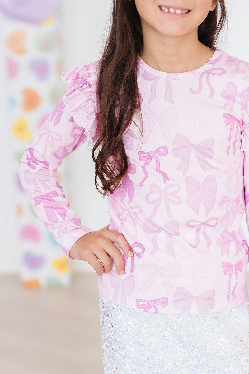 Twinkle Ties L/S Ruffle Tee styled by a smiling girl, featuring playful pink bow patterns and ruffled sleeves.