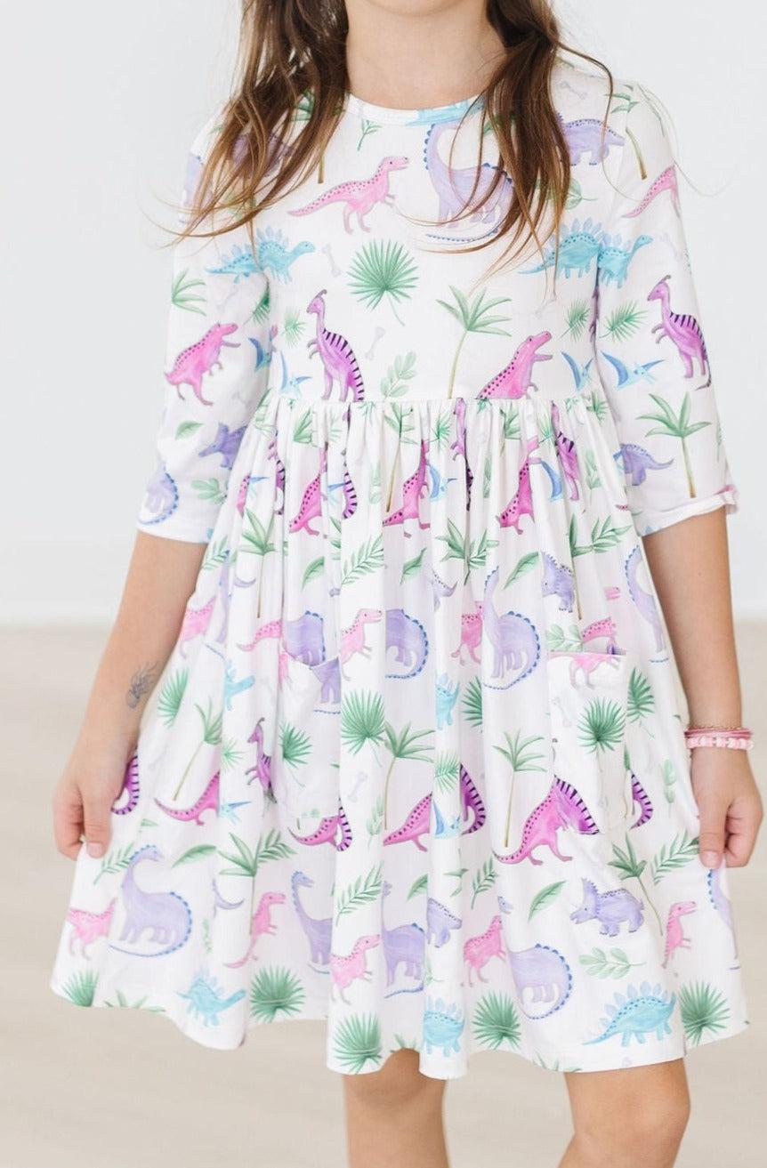 A joyful child wearing the Dino Darling 3/4 Sleeve Pocket Twirl Dress featuring a colorful dinosaur print, perfect for playtime and twirling.