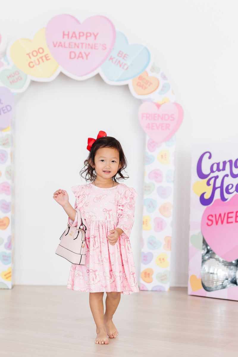 A little girl confidently strutting in the Howdy 3/4 Sleeve Ruffle Twirl Dress, holding a stylish handbag against a playful backdrop of colorful hearts.