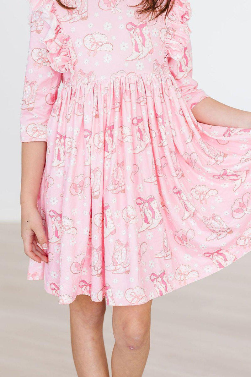 A young girl twirling joyfully in the Howdy 3/4 Sleeve Ruffle Twirl Dress, showcasing its playful pink fabric with charming designs of bows and flowers.
