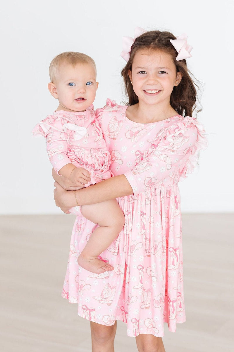 Two adorable siblings, a young girl and a baby, both dressed in matching Howdy 3/4 Sleeve Ruffle Twirl Dresses, bringing a burst of charm to their playful moment together.