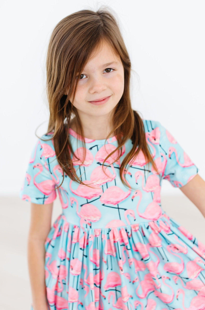 A close-up of a girl smiling softly in the Pretty in Pink Flamingos S/S Pocket Twirl Dress, emphasizing the playful flamingo graphics.