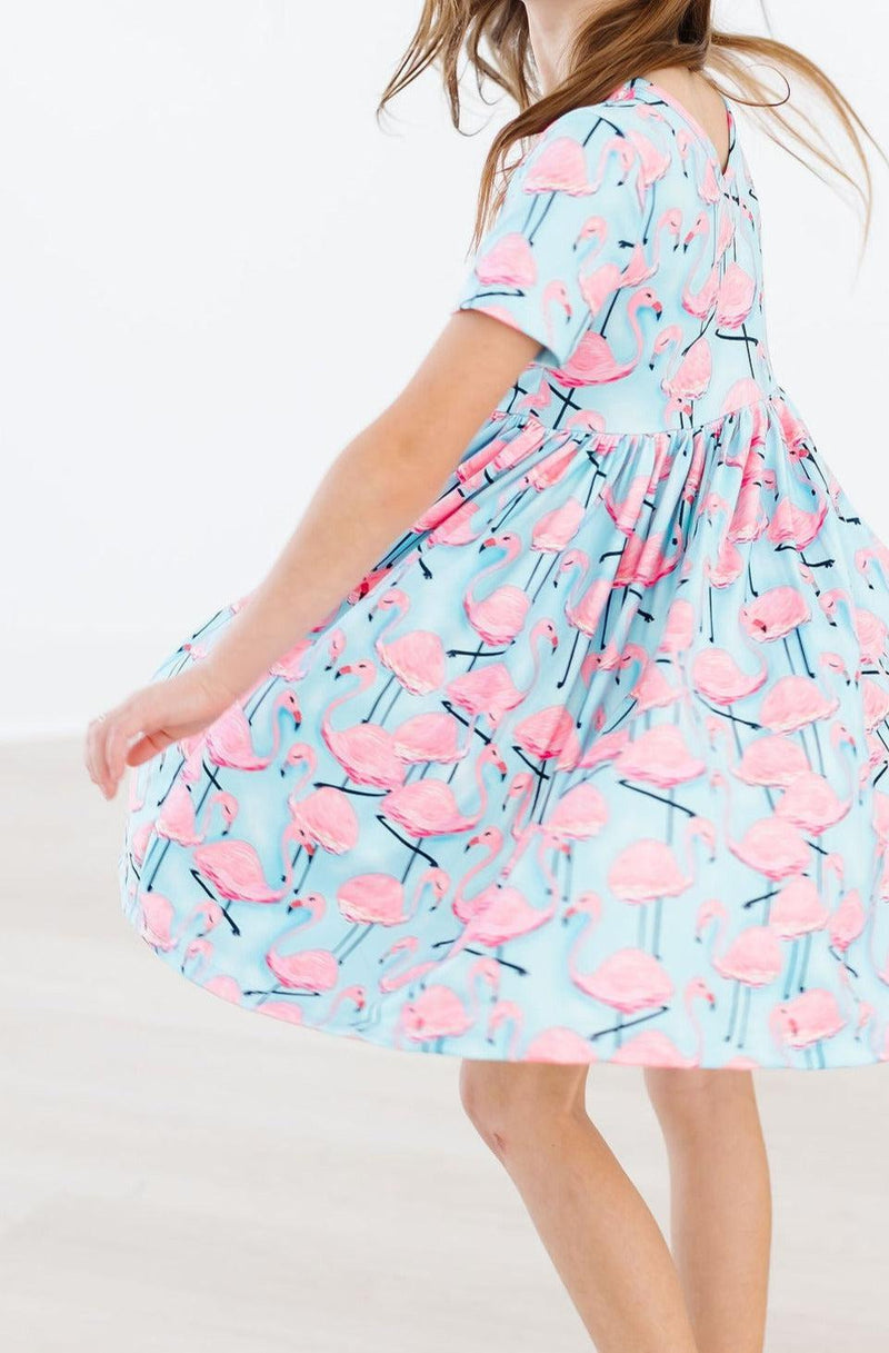 A joyful girl twirling in the Pretty in Pink Flamingos S/S Pocket Twirl Dress, her skirt flaring out in a burst of fun and color.