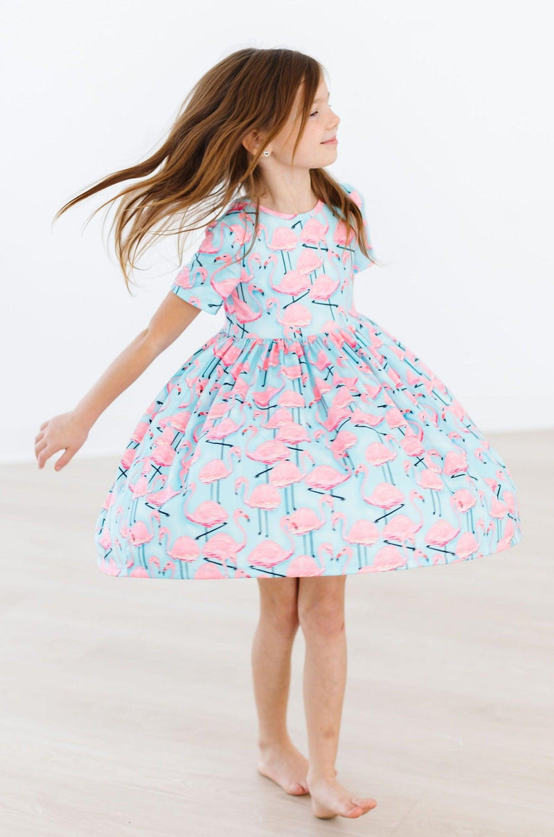 A girl elegantly twirling in the Pretty in Pink Flamingos S/S Pocket Twirl Dress, radiating fun and fashion with its vibrant print and carefree design.