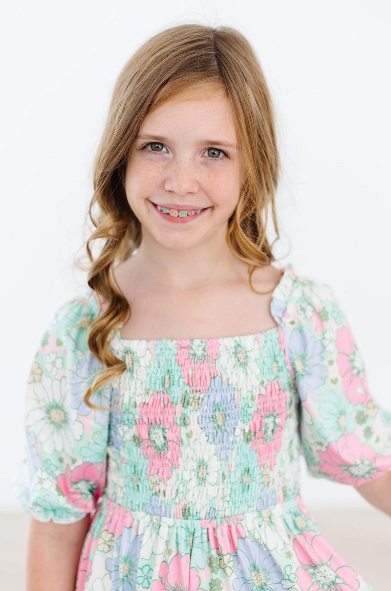 Smiling girl wearing the Minty Meadow Smocked Ruffle Dress, showcasing a lovely floral pattern with smocked bodice and playful ruffles.