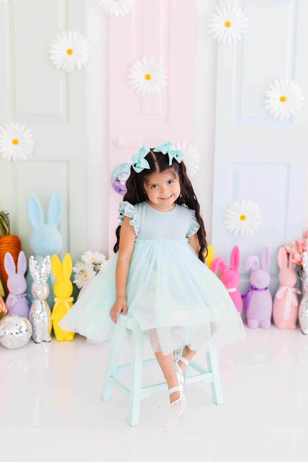 A young girl wearing a Sage Shimmer Tutu Dress from Mila & Rose, sitting on a teal stool against a pastel backdrop with decorative flowers and bunny figures, showcasing her playful style.