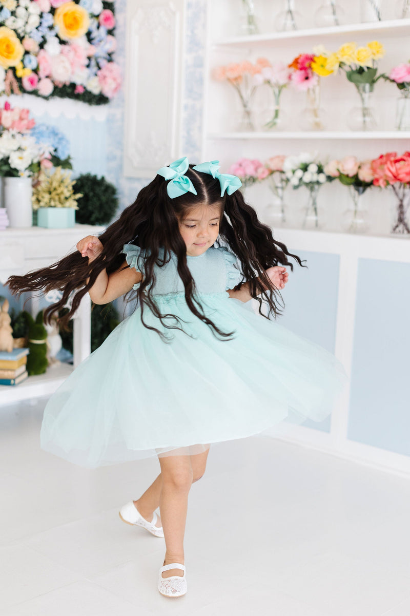 A girl in a Sage Shimmer Tutu Dress from Mila & Rose joyfully twirling, with her long hair flowing and bright flowers and decorations in the background, capturing the essence of fun.