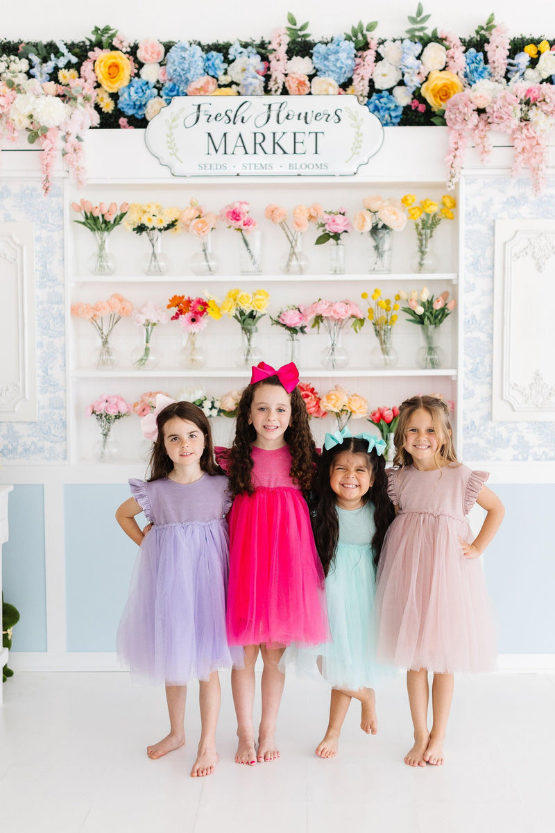 A delightful group of four girls proudly displaying their vibrant party outfits, with two in Lavender Shimmer Tutu Dresses, celebrating friendship and fun in a colorful flower market backdrop.