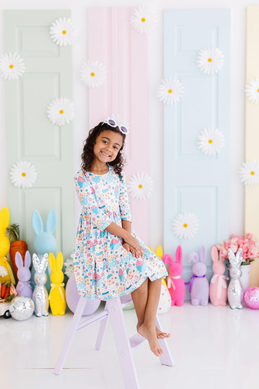 A joyful child sits on a stool, showcasing the Egg-cellent 3/4 Sleeve Pocket Twirl Dress, surrounded by pastel-colored decorations and playful bunny figures.
