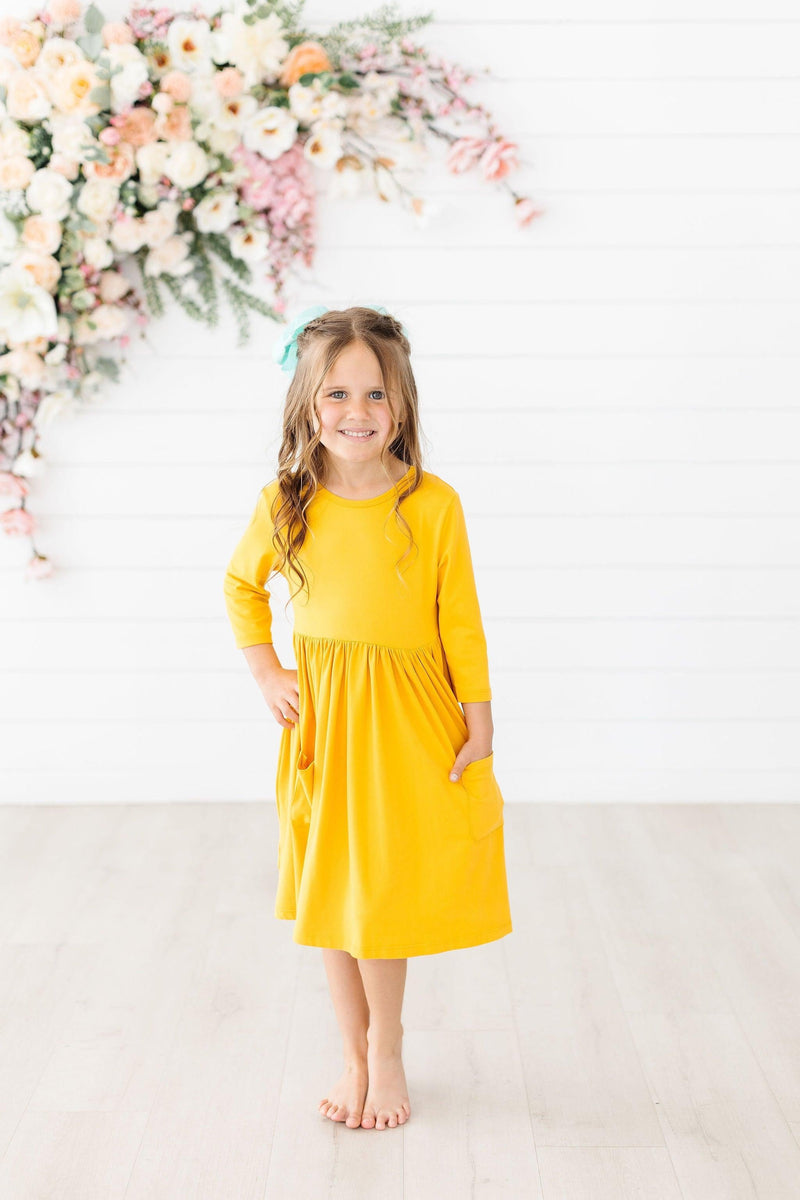 Girl posing in the Mustard 3/4 Sleeve Pocket Twirl Dress by Mila & Rose®, highlighting its pocket detail and sunny mustard color.