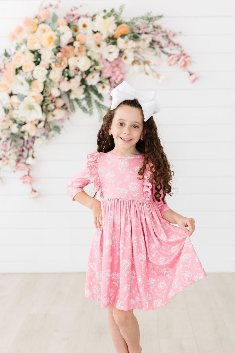 A young girl happily posing in the Dahlia Doodles 3/4 Sleeve Ruffle Twirl Dress, twirling in pink floral fabric, with a large bow in her hair and a floral backdrop.