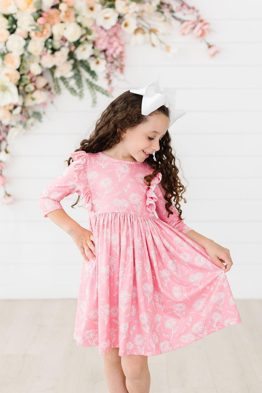 A young girl wearing the Dahlia Doodles 3/4 Sleeve Ruffle Twirl Dress by Mila & Rose, showcasing its playful design with ruffle details and floral patterns, perfect for twirling and everyday adventures.