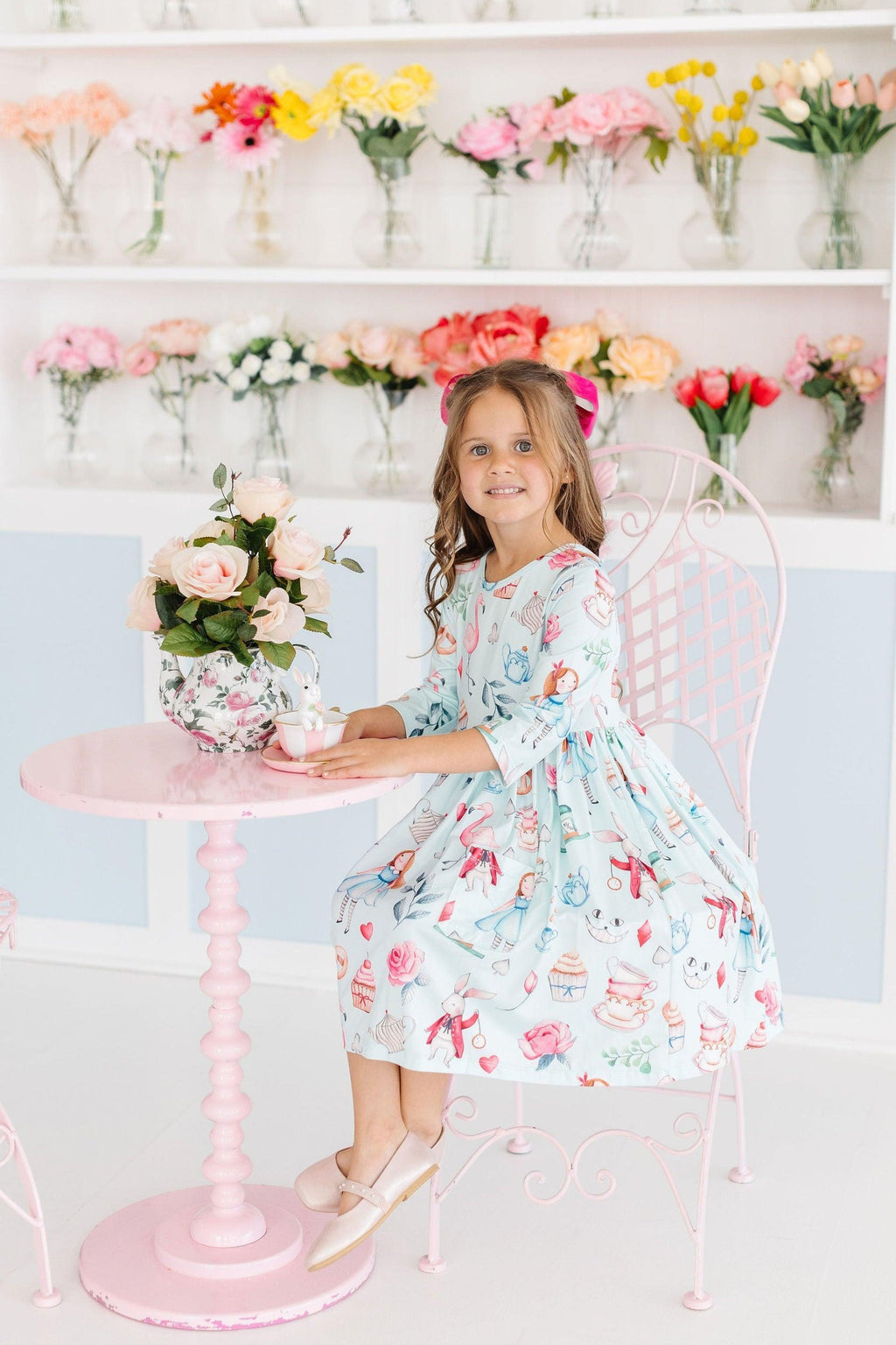 A girl sitting at a pink table wearing the Wonderland 3/4 Sleeve Pocket Twirl Dress, surrounded by colorful flowers in vases, showcasing the dress in a charming tea party setting.
