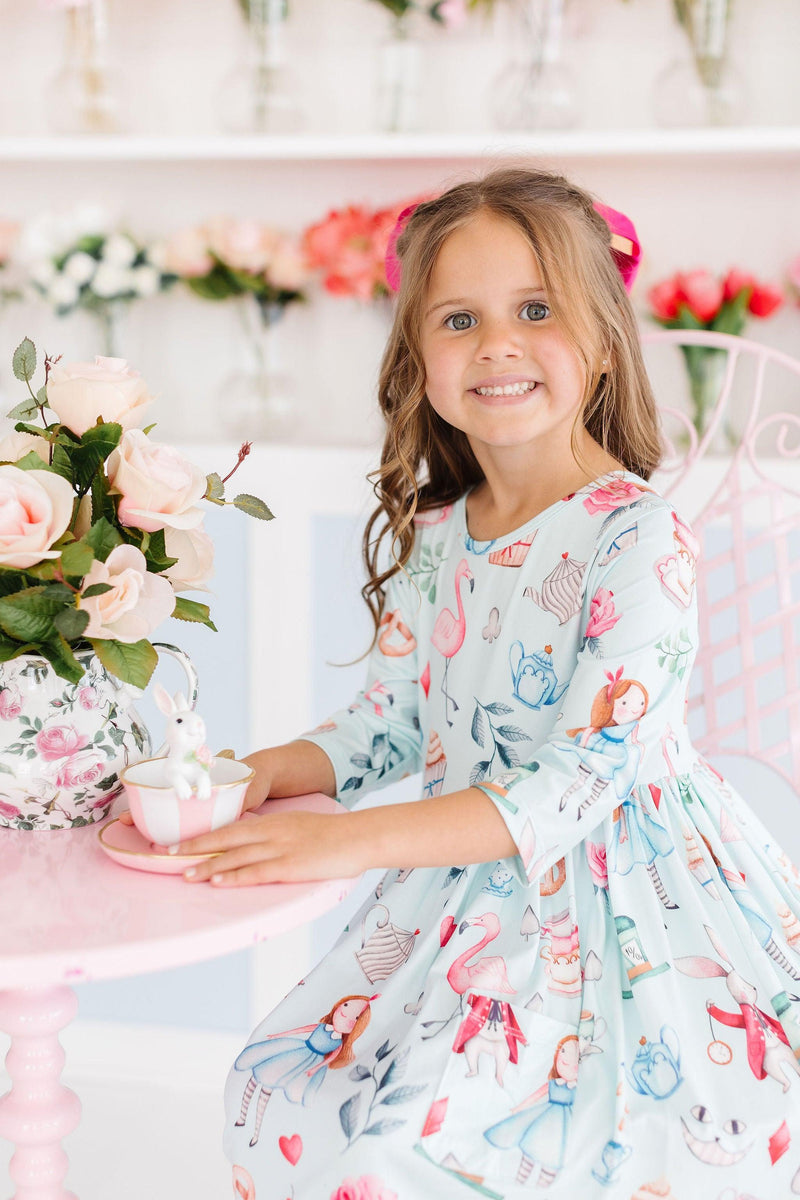 A girl in the Wonderland 3/4 Sleeve Pocket Twirl Dress sitting at a pink table with a floral teapot, smiling and enjoying a delightful tea party atmosphere.