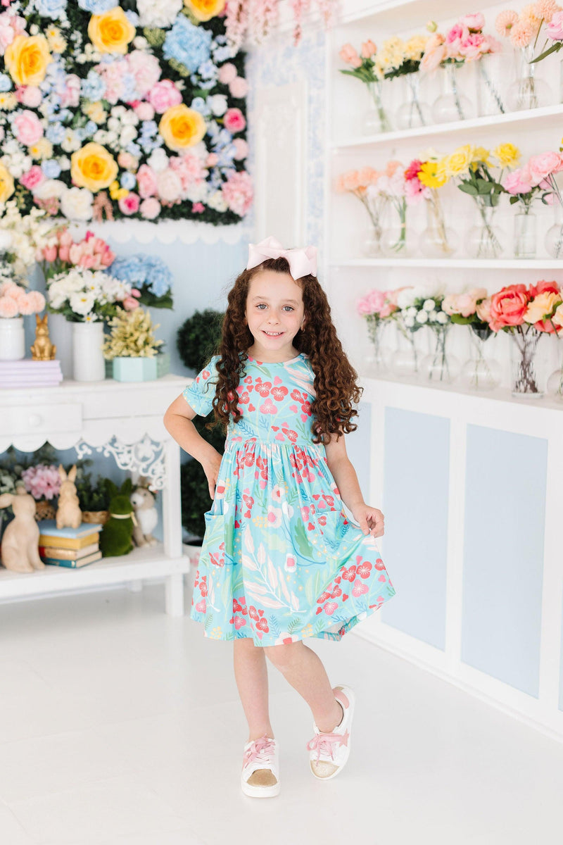 A young girl strikes a confident pose in the Spring Breeze S/S Pocket Twirl Dress, surrounded by a brightly decorated floral backdrop.