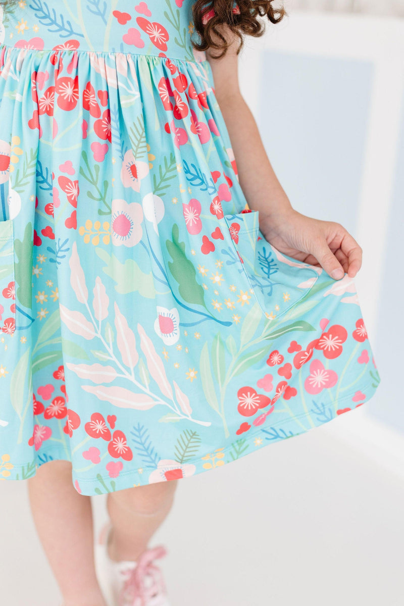 A young girl highlights the pocket detail of the Spring Breeze S/S Pocket Twirl Dress while twirling, showcasing the playful floral design.