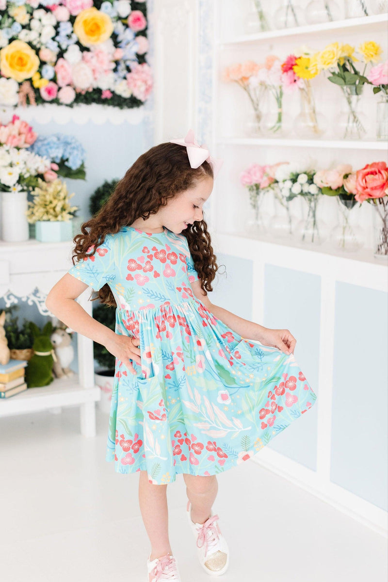 A smiling girl in a stylish Spring Breeze S/S Pocket Twirl Dress poses playfully, displaying the beautiful floral pattern in a colorful room.