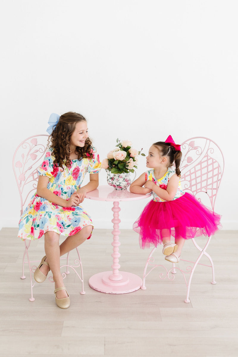 Two girls in Colorful Rosie Floral Smocked Ruffle Dresses enjoying a cheerful moment at a pink table, highlighting the joyful spirit of the smocked ruffle dress.