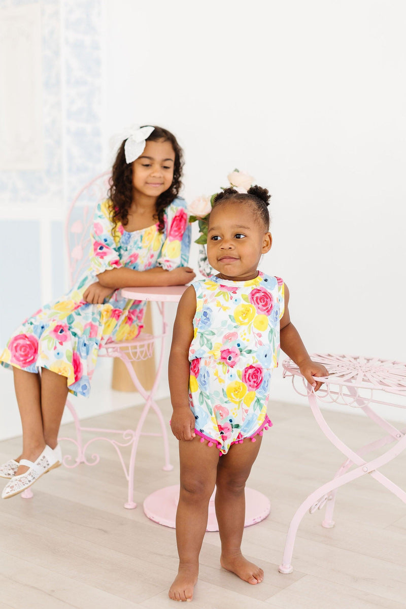 Sisters wearing Colorful Rosie Floral Smocked Ruffle Dresses and a playful romper, emphasizing the fun and style of the smocked ruffle dress collection.