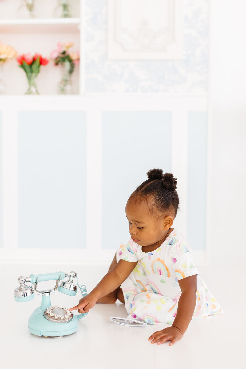 Watercolor Rainbows S/S Twirl Bodysuit worn by a cute child sitting on the floor, engaged with a retro phone toy, embodying fun and playfulness.