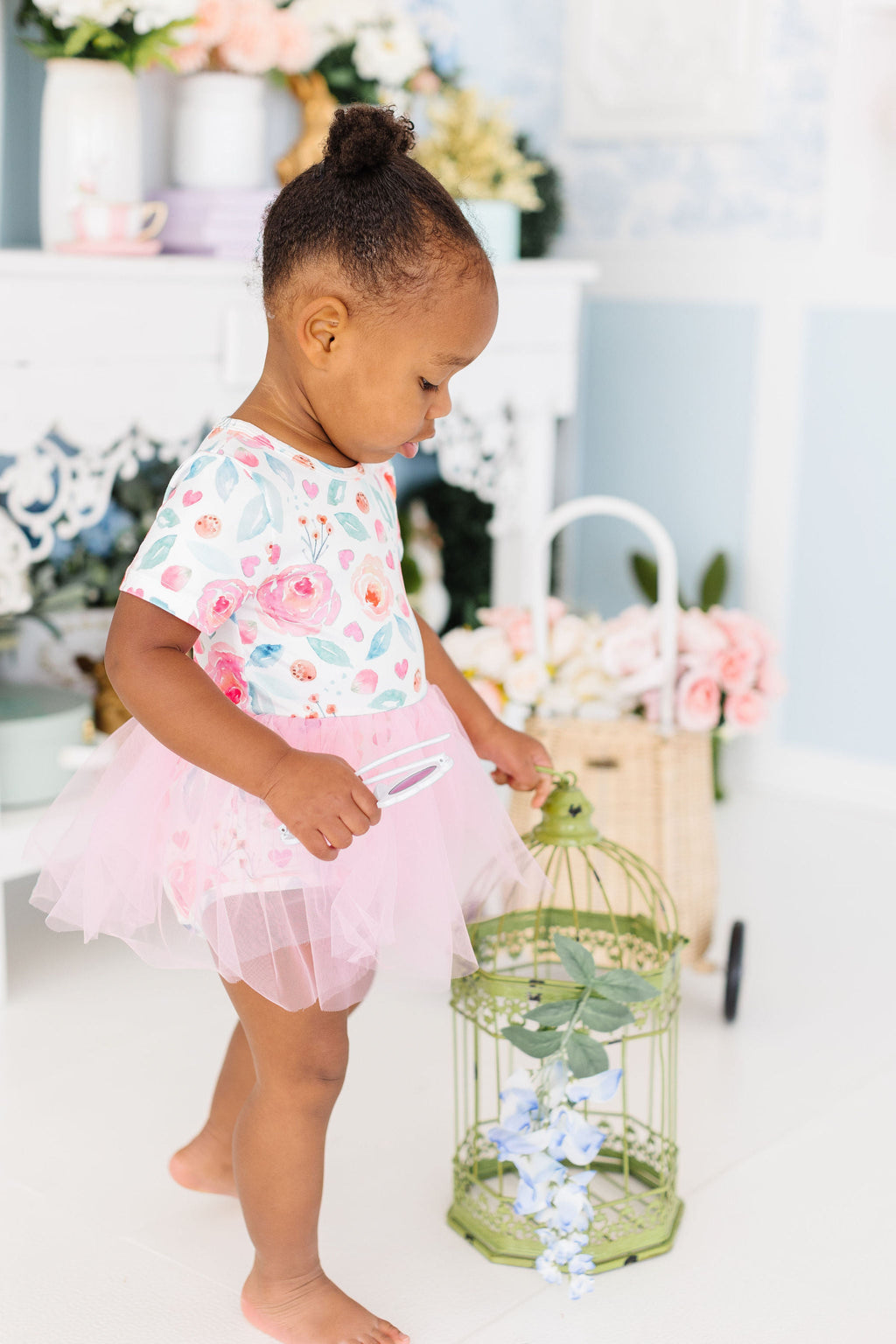 Baby girl in a SALE Rosettes Pink Tutu Bodysuit for Babies and Toddlers, playing with a decorative cage in a bright, flower-adorned room.