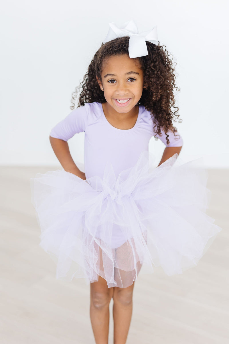 Little girl happily modeling the Lavender 3/4 Tutu Leotard with a fluffy tutu and a big white bow, showcasing her dance spirit with a beaming smile.