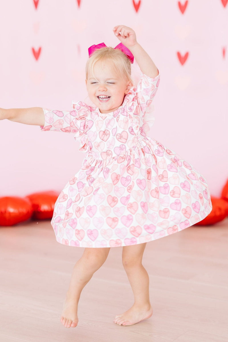 A young girl spinning happily in the Melt My Heart 3/4 Sleeve Ruffle Twirl Dress, radiating joy amidst playful heart decorations.