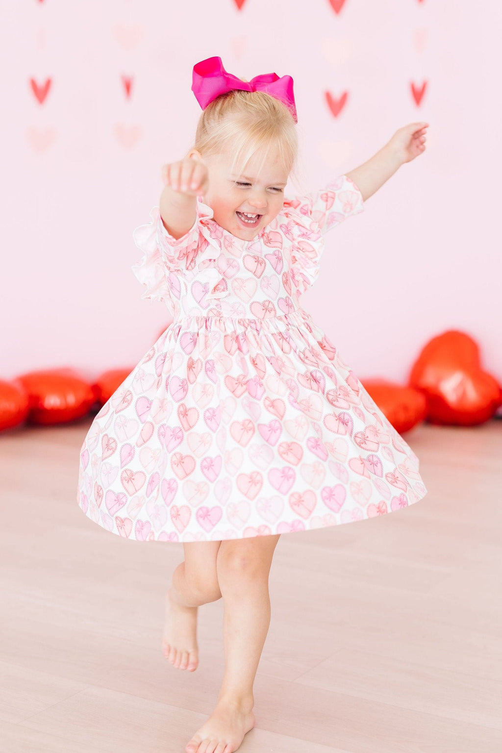 Adorable little girl twirling joyfully in the Melt My Heart 3/4 Sleeve Ruffle Twirl Dress, surrounded by festive heart decorations, showcasing fun and style in a vibrant pink atmosphere.