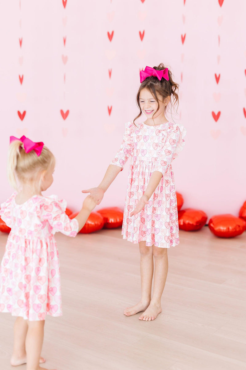 Two girls in heart-patterned Melt My Heart 3/4 Sleeve Ruffle Twirl Dress, enjoying playtime together in a pink-themed decor with heart-shaped decorations.