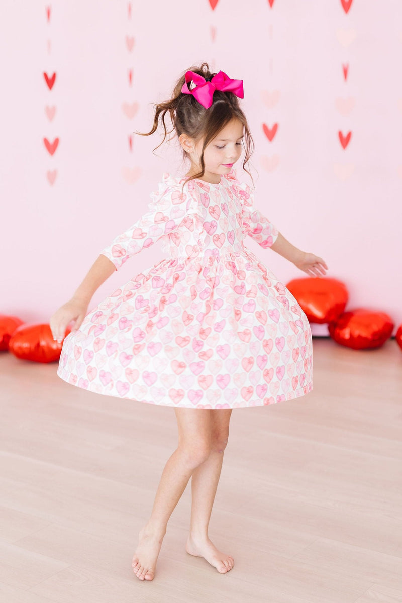 A girl twirling gracefully in the Melt My Heart 3/4 Sleeve Ruffle Twirl Dress, surrounded by colorful heart-shaped cushions in a whimsical setting.