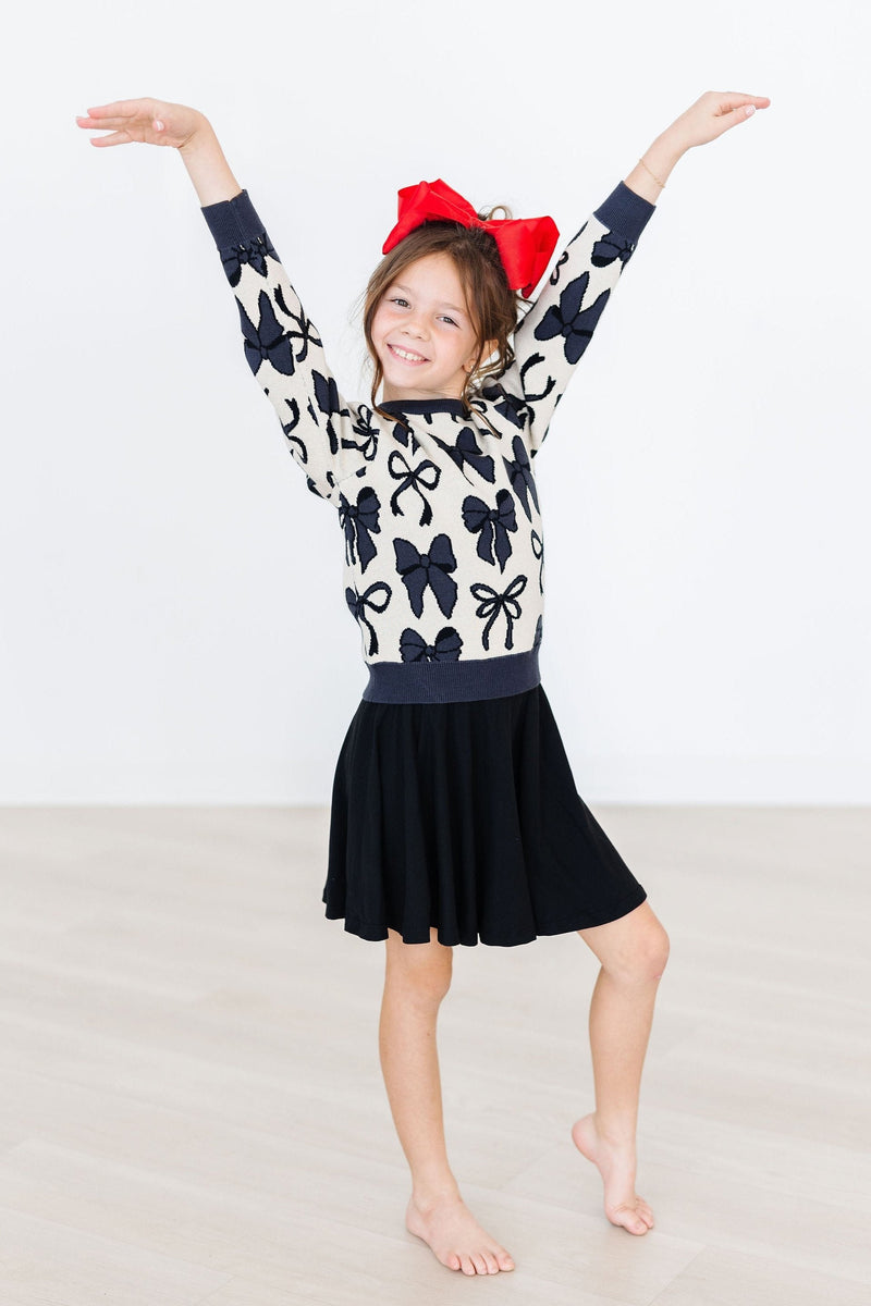 A cheerful girl posing with arms raised, confidently wearing a Black Twirl Skort by Mila & Rose, complemented by a fun bow-adorned sweater.