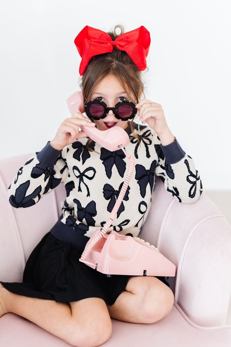A joyful girl wearing sunglasses and a red bow in her hair, posing with a pink retro phone while dressed in the Black Twirl Skort from Mila & Rose.