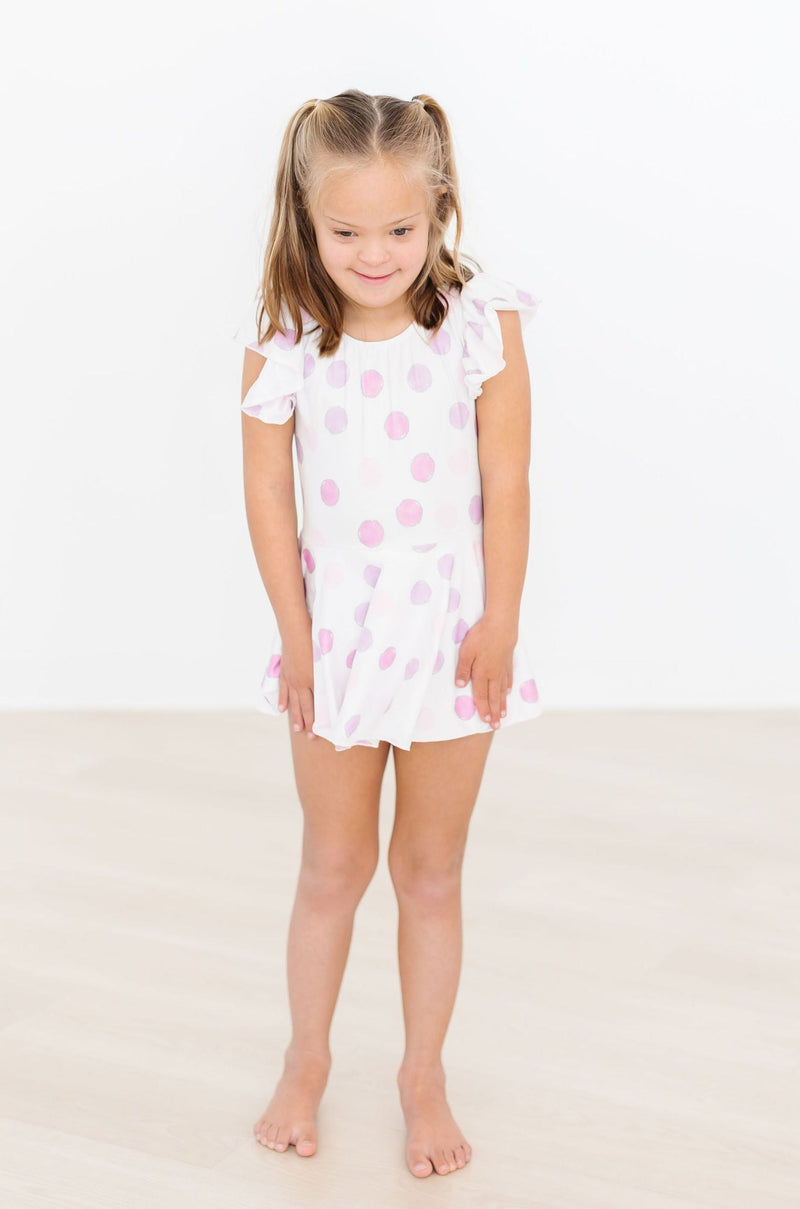 Little girl wearing the Pastel Polka Dot S/S Twirl Leotard, striking a cute pose with a smile, showcasing its fun design and flutter sleeves.