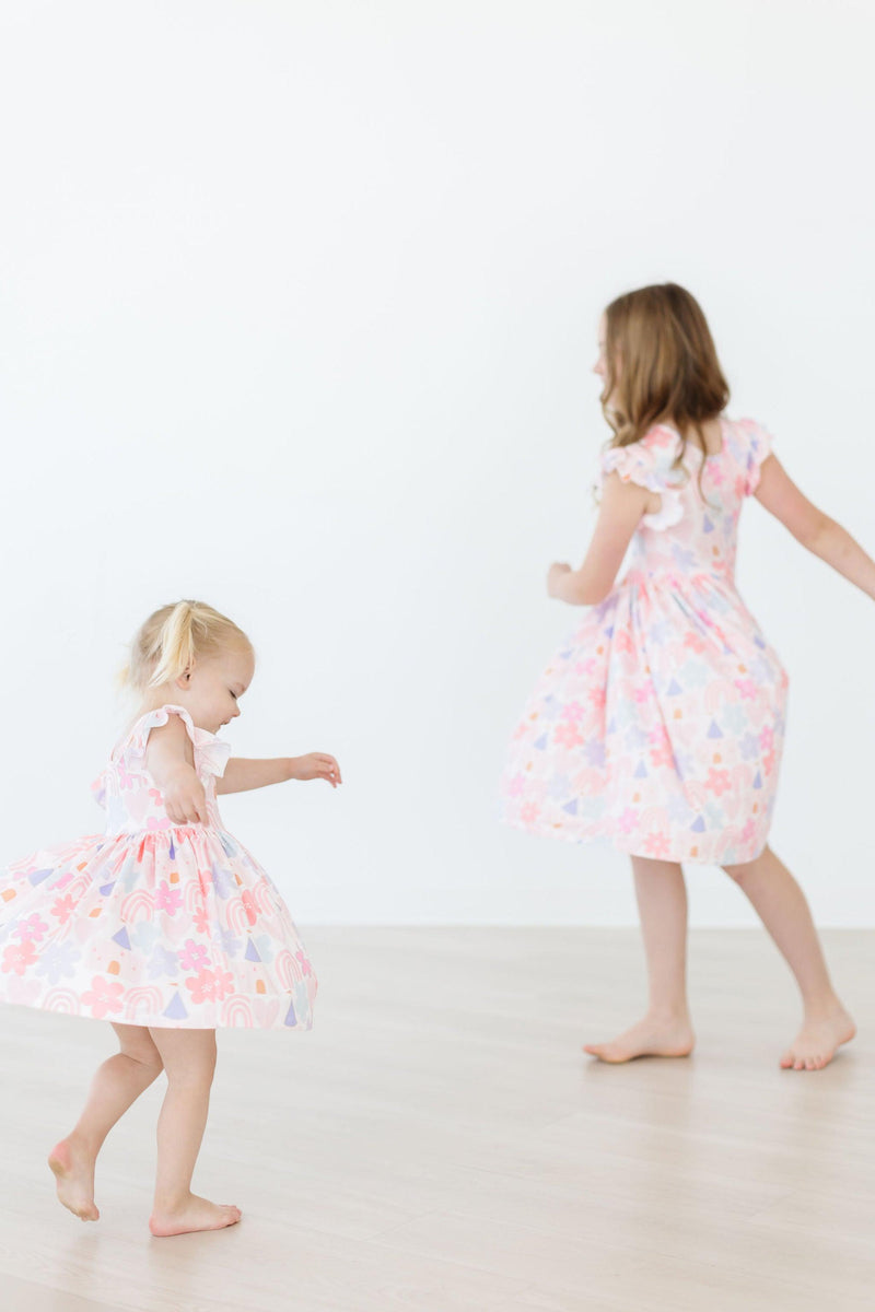 Little girl joyfully twirling in the Castles & Rainbows Flutter Sleeve Twirl Dress, showcasing colorful patterns and flutter sleeves.