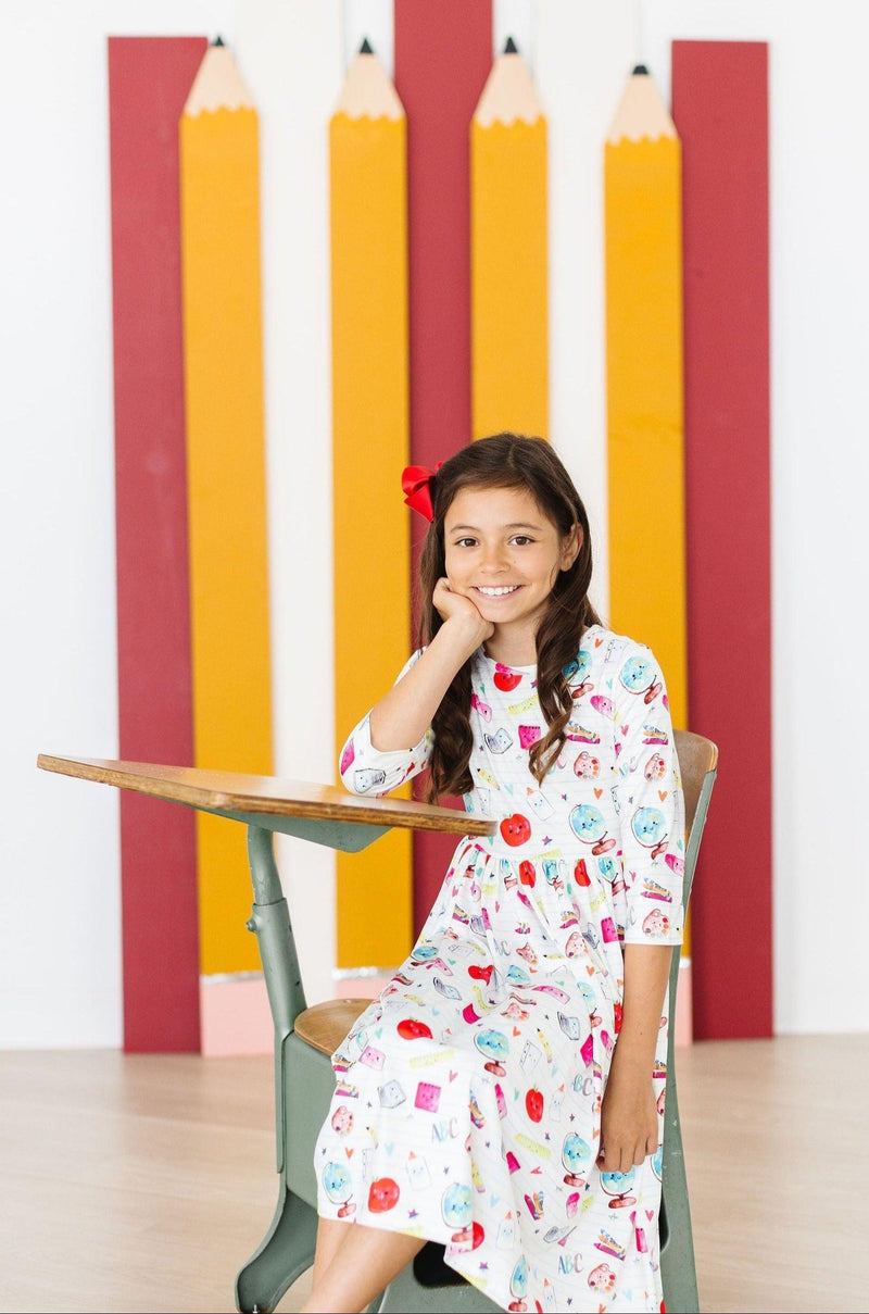 A girl seated at a desk, thoughtfully posing for the camera in the Smarty Pants Pocket Twirl Dress by Mila & Rose, with a colorful backdrop of painted pencil shapes.