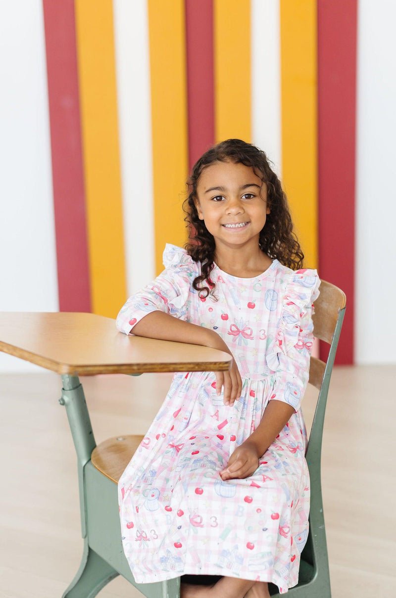 A girl seated at a desk, wearing the School Days 3/4 Ruffle Twirl Dress by Mila & Rose ®, smiling in a playful classroom environment.