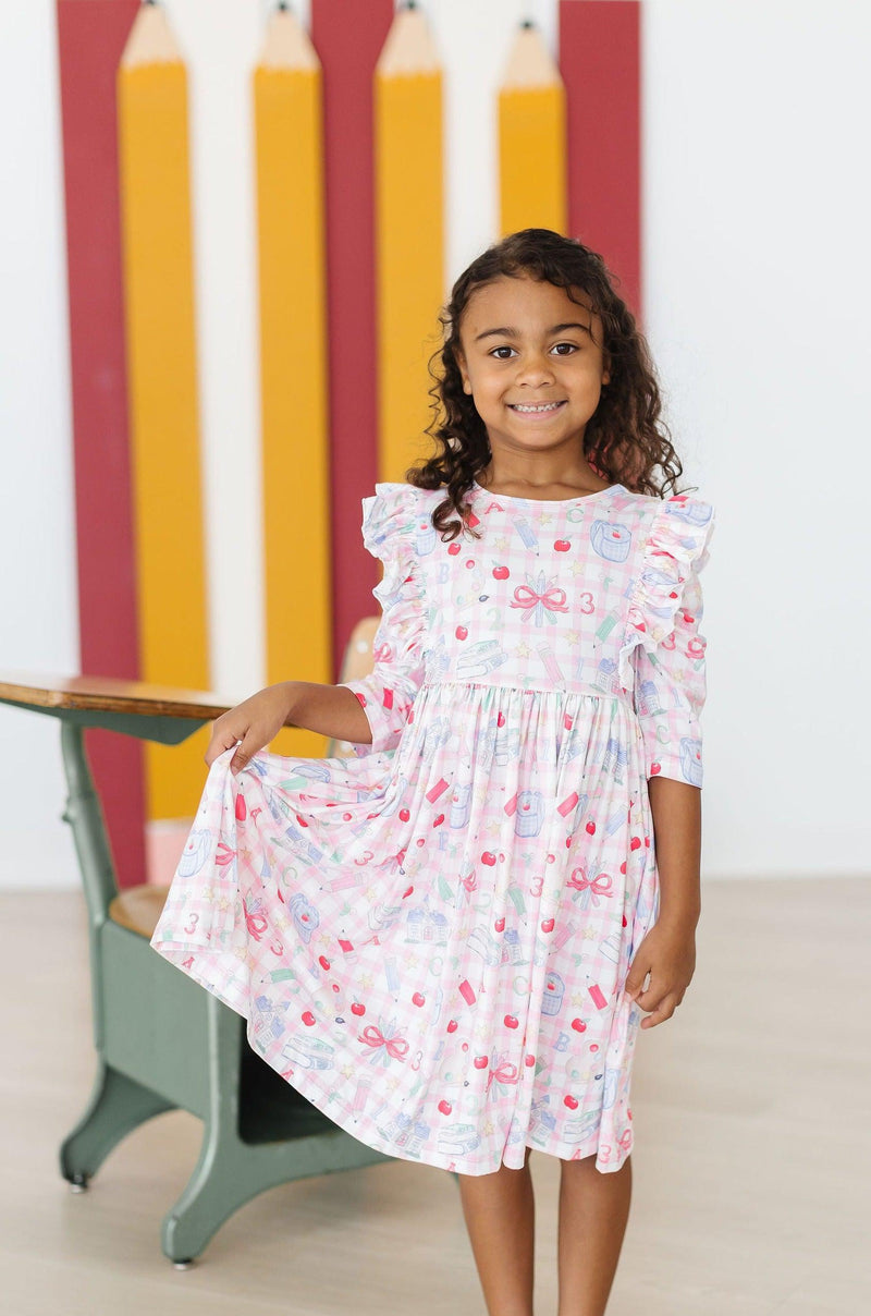 A girl smiling while wearing the School Days 3/4 Ruffle Twirl Dress by Mila & Rose ®, displaying playful patterns in a bright classroom.
