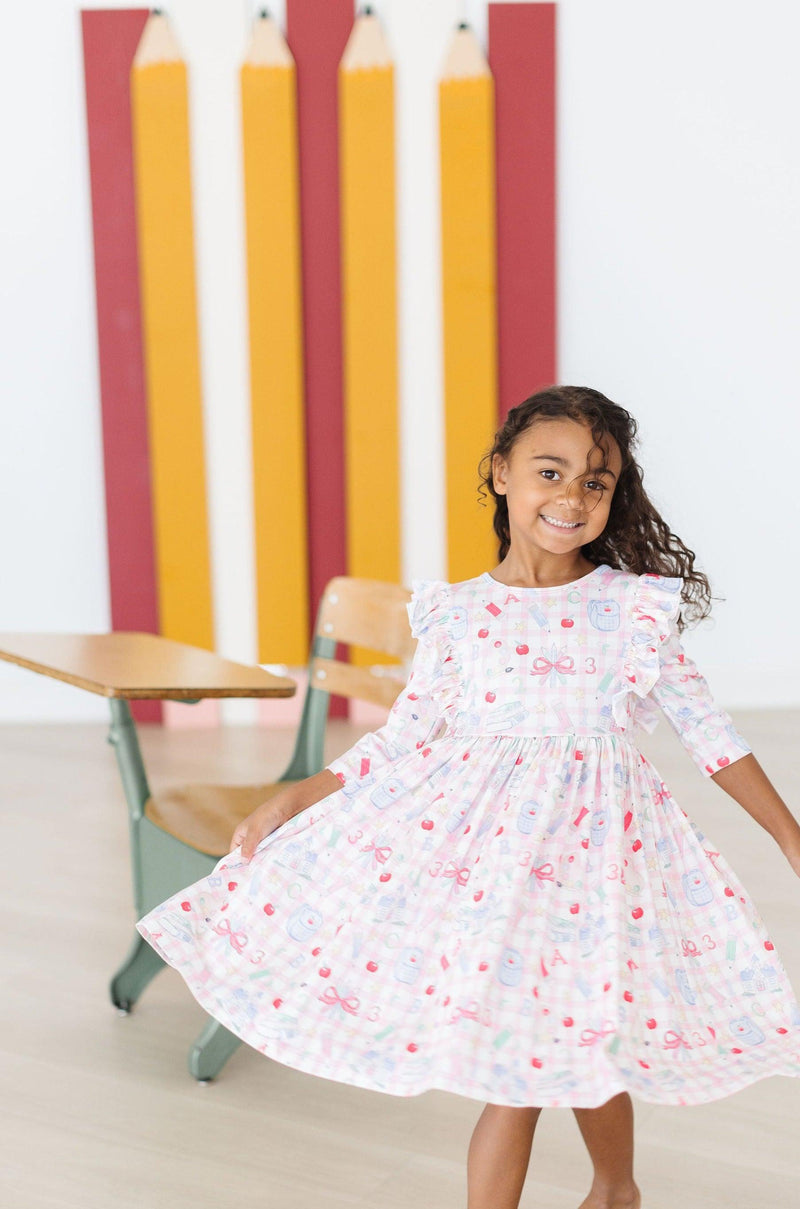 A girl twirling in her School Days 3/4 Ruffle Twirl Dress by Mila & Rose, showcasing a playful design with apples against a cheerful classroom backdrop, perfect for capturing joyful moments.