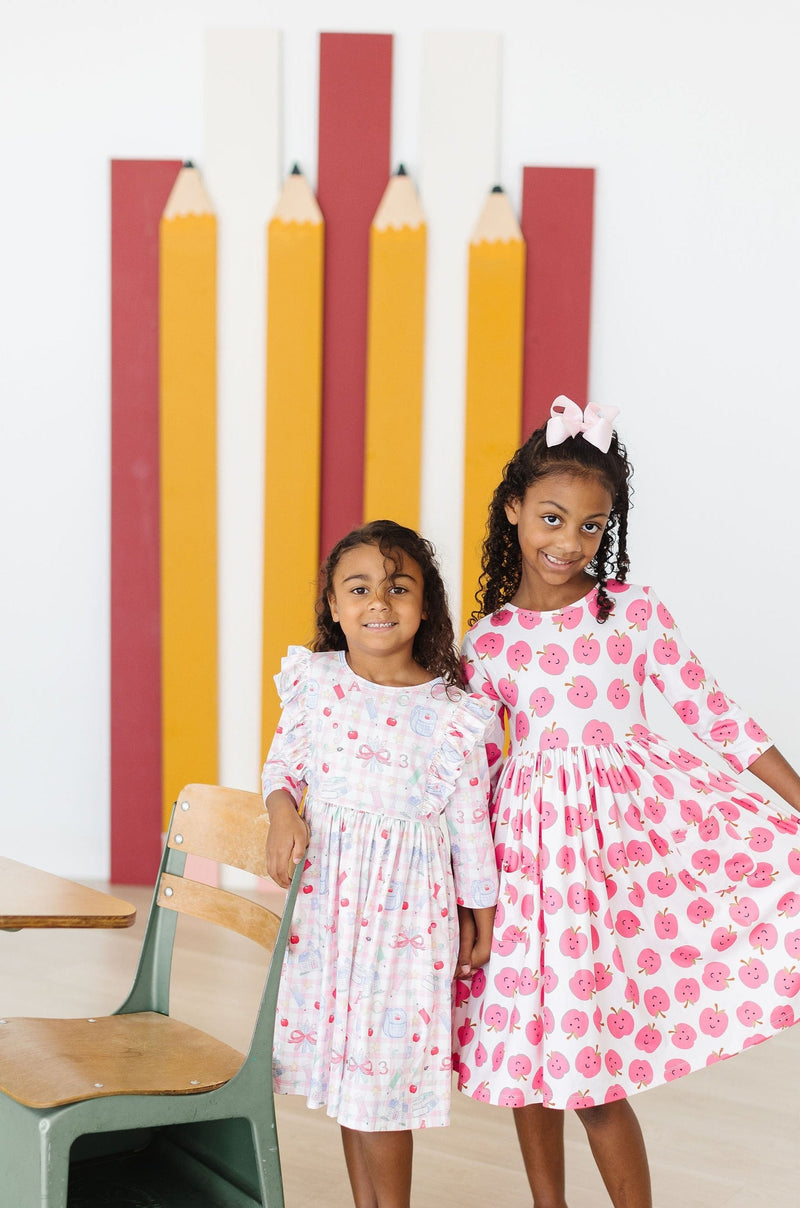 Two girls wearing the School Days 3/4 Ruffle Twirl Dress by Mila & Rose, one in a pink dress with apples, and the other in a light floral dress, happy and enjoying a fun moment in a colorful classroom setting.