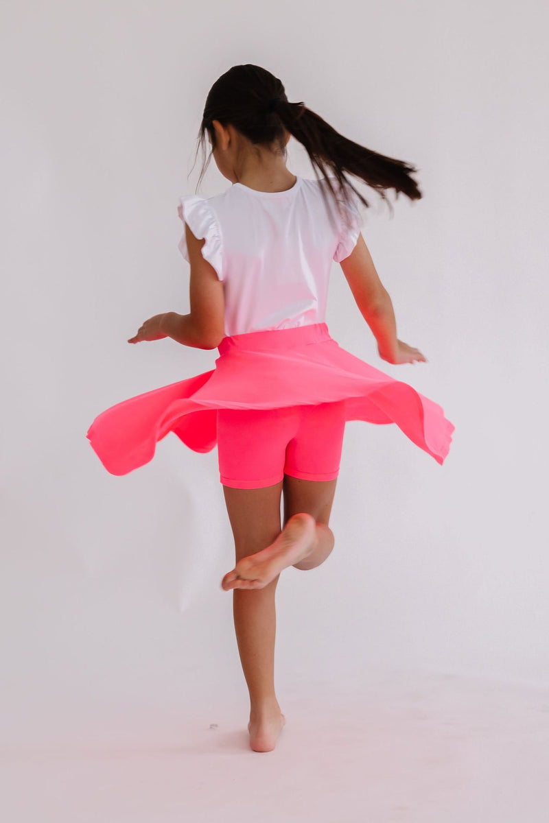 A girl joyfully twirling in a Neon Pink Twirl Skort from Mila & Rose, showcasing the vibrant skirt and attached shorts as she spins barefoot against a plain background.