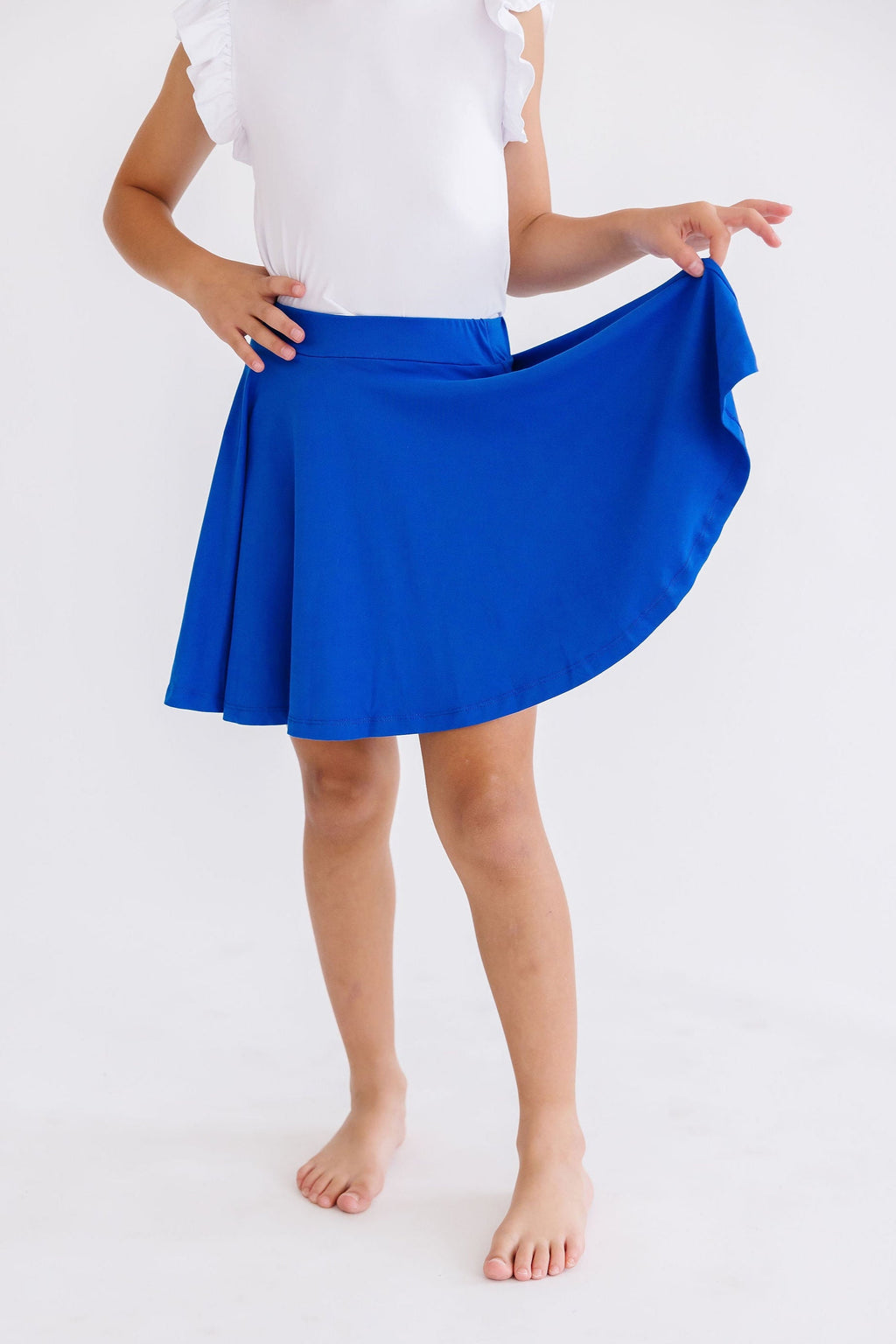 A young girl wearing a Royal Blue Twirl Skort from Mila & Rose, playfully lifting the hem of the skort to showcase its twirl effect.