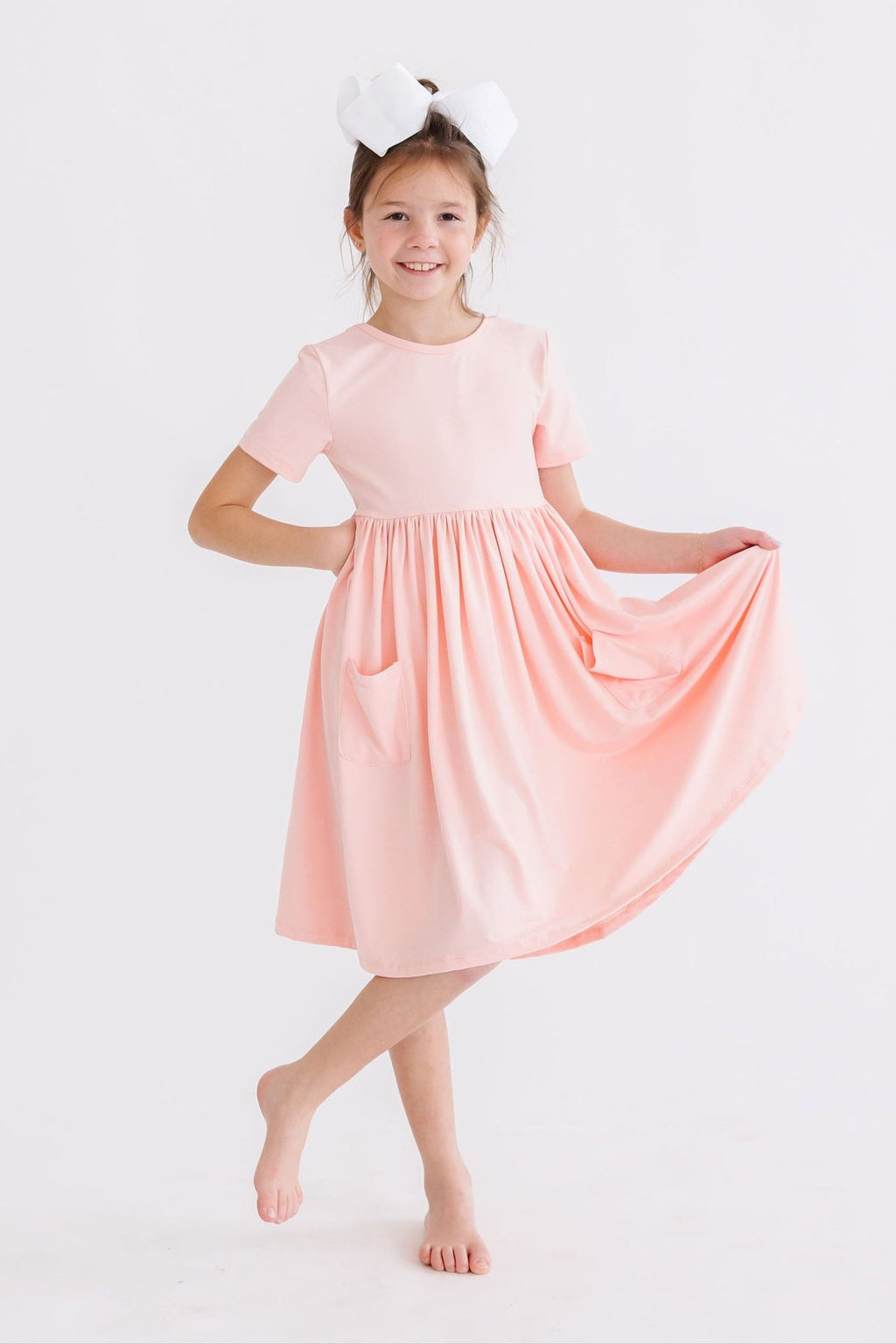 A joyful girl in a Peach S/S Pocket Twirl Dress by Mila & Rose, dancing with her dress flowing, showcasing its playful pockets.