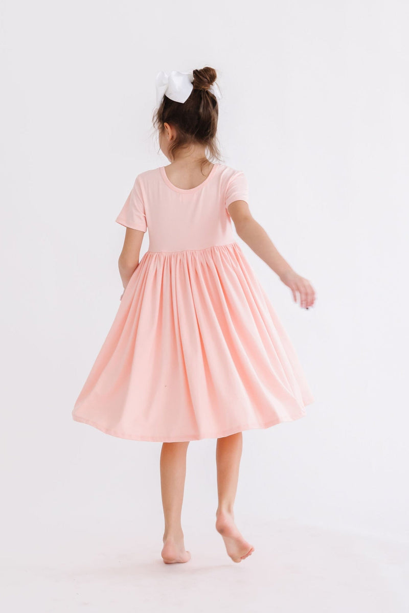 Back view of a girl twirling in a Peach S/S Pocket Twirl Dress by Mila & Rose, highlighting the dress's flared skirt and scoop back design.