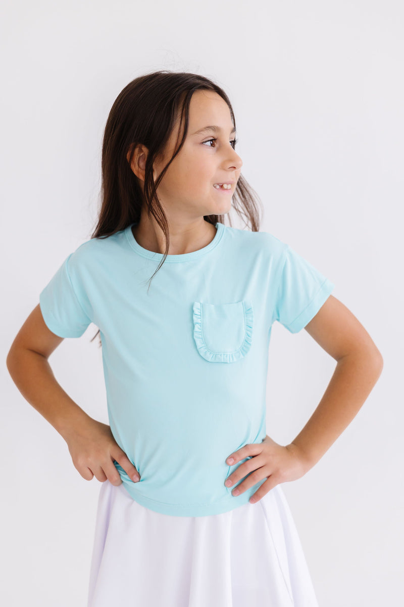 Delightful Aqua Ruffle Pocket Tee by Mila & Rose, featuring a charming young girl with a bright smile, striking a pose while displaying the stylish ruffled pocket tee, ideal for any fashionable occasion.